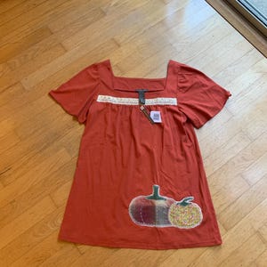 May include: Rust-colored short-sleeve dress with a square neckline and a white lace trim. The dress features two pumpkin appliqués on the front. A tag is attached near the neckline. The dress is made of a soft material.