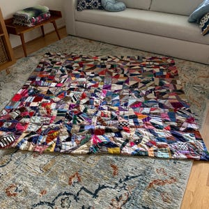 May include: A vibrant patchwork quilt with a geometric design, showcasing diverse fabric patterns and colours such as red, blue, and yellow. The quilt is displayed on a patterned rug in a living room.