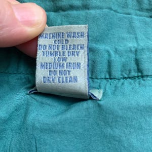 Pu&ograve; includere: Primo piano di un tessuto verde acqua con un'etichetta di cura. L'etichetta, con testo blu, recita "MACHINE WASH COLD, DO NOT BLEACH, TUMBLE DRY LOW, MEDIUM IRON, DO NOT DRY CLEAN."
