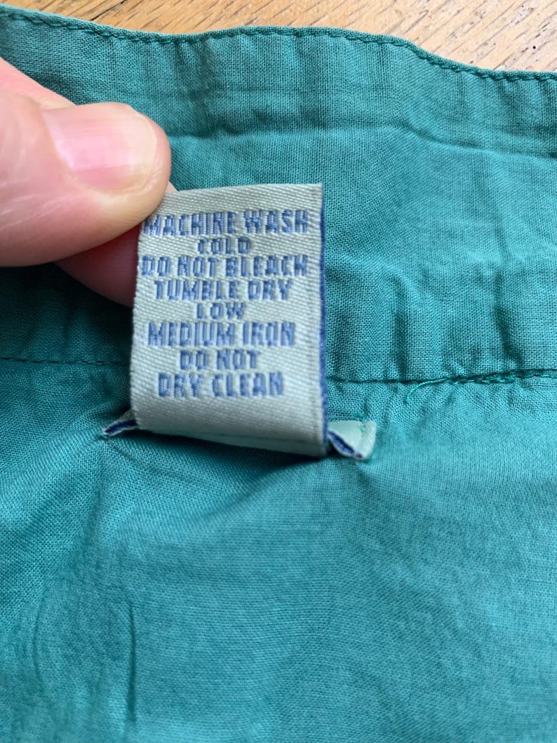 Pu&ograve; includere: Primo piano di un tessuto verde acqua con un'etichetta di cura. L'etichetta, con testo blu, recita "MACHINE WASH COLD, DO NOT BLEACH, TUMBLE DRY LOW, MEDIUM IRON, DO NOT DRY CLEAN."