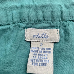 Pu&ograve; includere: Primo piano di un capo color turchese con etichetta. L'etichetta dice "odille" in corsivo, con testo sottostante che indica che il capo &egrave; 100% cotone, prodotto in India e taglia 2. Testo aggiuntivo: RN e CA.