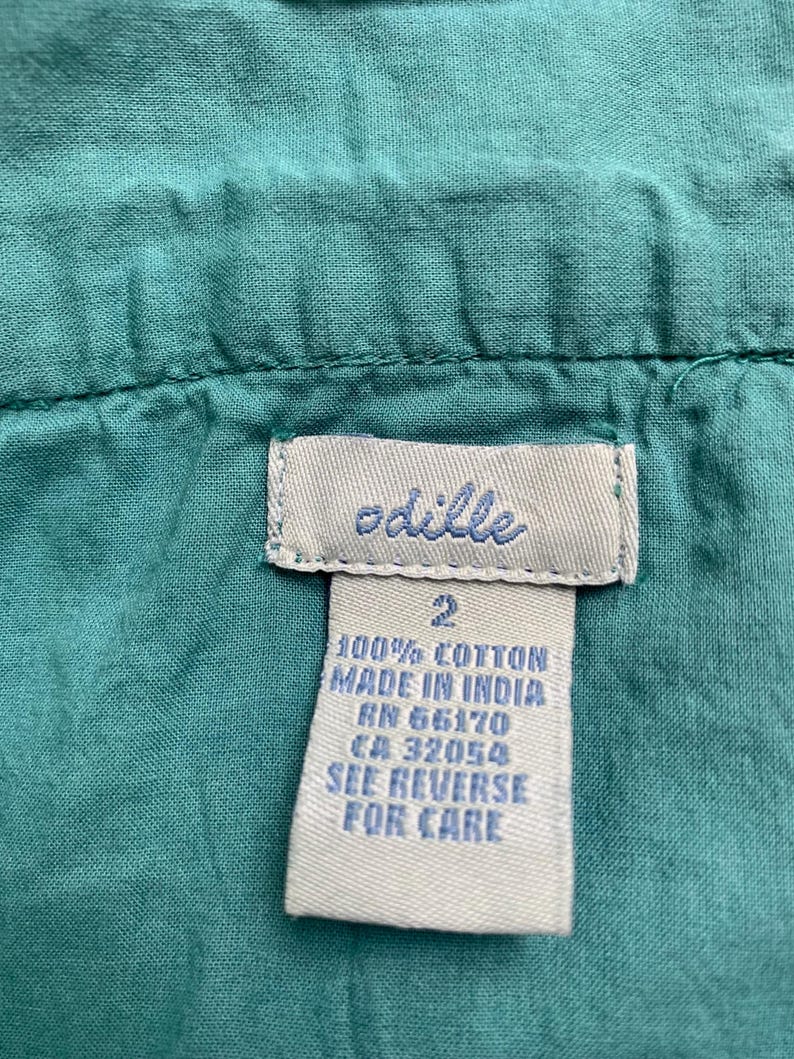 Pu&ograve; includere: Primo piano di un capo color turchese con etichetta. L'etichetta dice "odille" in corsivo, con testo sottostante che indica che il capo &egrave; 100% cotone, prodotto in India e taglia 2. Testo aggiuntivo: RN e CA.
