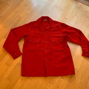 May include: A bright red button-down shirt with two chest pockets. The long-sleeved shirt is made of a soft material and has a classic collar. The shirt is laid flat on a wooden surface.
