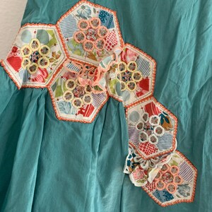 Pu&ograve; includere: Un capo color turchese con un design patchwork decorativo. Il patchwork presenta forme esagonali con tessuto floreale e fantasia, accentuato da finiture arancioni e dettagli ricamati.
