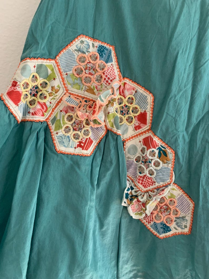 Pu&ograve; includere: Un capo color turchese con un design patchwork decorativo. Il patchwork presenta forme esagonali con tessuto floreale e fantasia, accentuato da finiture arancioni e dettagli ricamati.