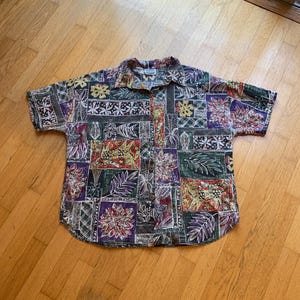 Puede incluir: Una camisa de manga corta con botones y un estampado de patchwork multicolor que presenta varios motivos tropicales, incluyendo flores estilizadas, hojas y figuras de animales en tonos morado, verde, amarillo y rojo sobre un fondo oscuro.