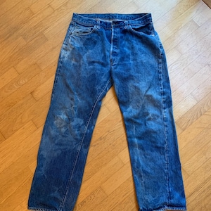 May include: Faded blue denim jeans with a button fly. The jeans show signs of wear and tear, with lighter patches and a classic five-pocket design. The straight-leg cut is visible.