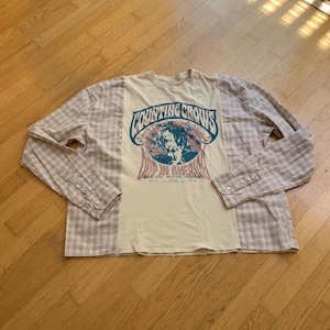 Peut inclure: Une chemise à manches longues avec un panneau central crème arborant une impression graphique du groupe Counting Crows et le texte "Hard Candy in America". Les manches et les panneaux latéraux sont en tissu à carreaux rose clair et blanc.
