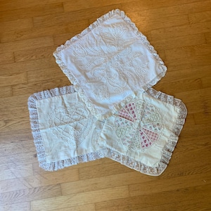 May include: Three off-white decorative pillow covers with lace trim. One pillow cover features a floral design, another a heart, and the third a patchwork fan design. The pillows are arranged on a wooden floor.