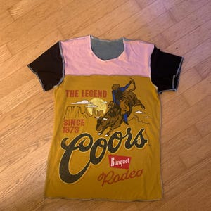以下が含まれることがあります： ロデオのデザインと「Coors Banquet Rodeo」のテキストが特徴のマスタードイエローのフロントパネルが付いたパッチワークTシャツ。トップはピンクのネックライン、ブルーのヨーク、ブラックのスリーブです。デザインには、雄牛に乗るカウボーイが含まれています。