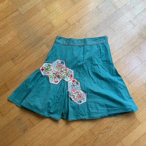 Pu&ograve; includere: Gonna color verde acqua con un design patchwork unico. La gonna presenta una serie di toppe esagonali in vari colori e motivi, aggiungendo un tocco di fantasia. Ha una silhouette svasata e un bordo arancione a contrasto in vita.