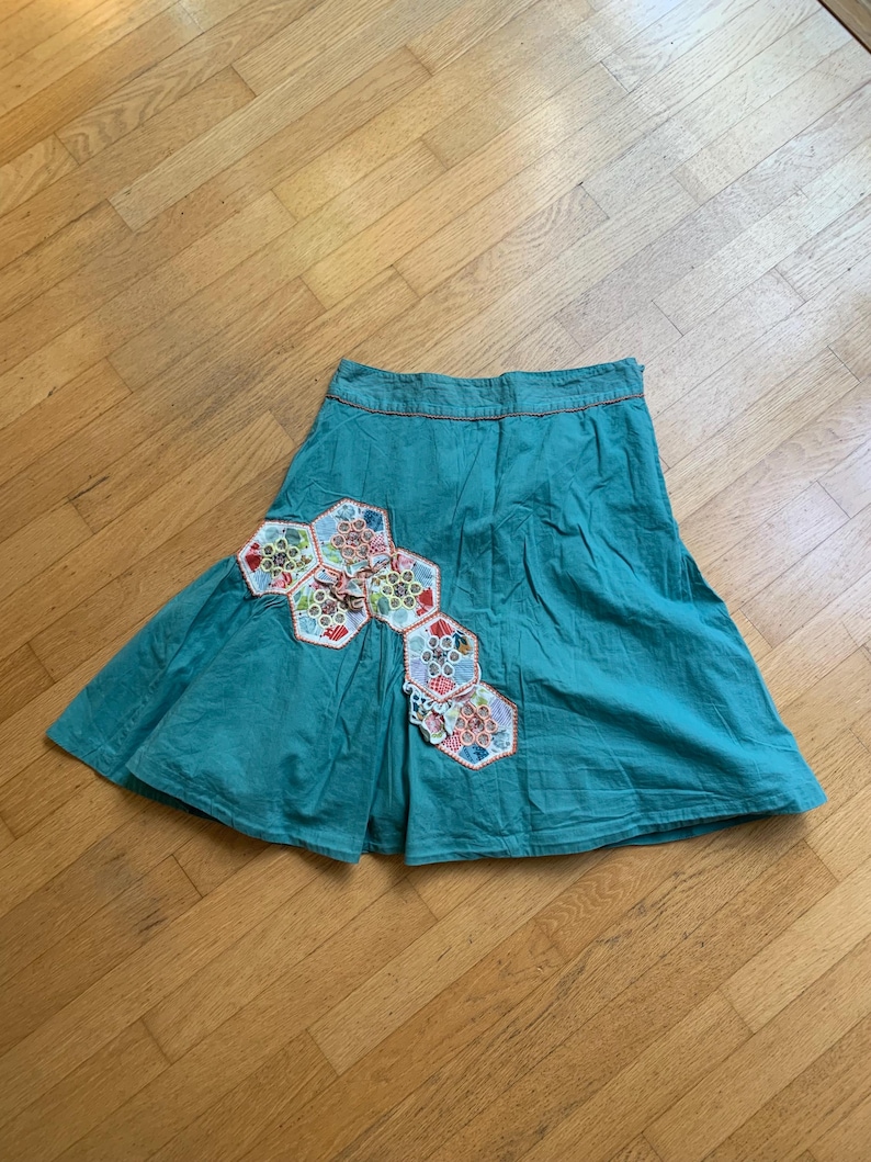 Pu&ograve; includere: Gonna color verde acqua con un design patchwork unico. La gonna presenta una serie di toppe esagonali in vari colori e motivi, aggiungendo un tocco di fantasia. Ha una silhouette svasata e un bordo arancione a contrasto in vita.