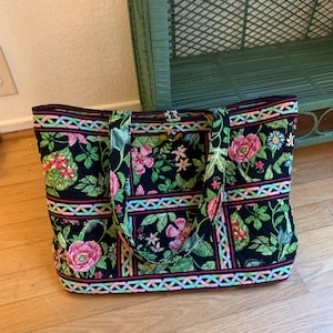 Puede incluir: Bolso tote negro con un estampado floral de flores y hojas rosas y verdes. El bolso es rectangular con dos asas y un ribete en rosa, azul y verde. Adecuado para llevar objetos.