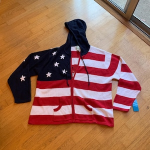 May include: A knit zip-up hoodie featuring a patriotic design with a navy blue upper section adorned with white stars and a lower section with horizontal red and white stripes. The garment includes a hood with drawstrings and two front pockets.