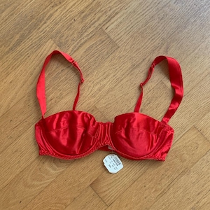 Pode incluir: Um sutiã com aros de cetim vermelho brilhante com alças ajustáveis e uma etiqueta branca de venda presa ao centro. A etiqueta exibe o tamanho 34A e um código de barras. O sutiã apresenta um acabamento liso e brilhante.