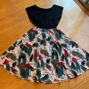 May include: A dress featuring a solid black bodice with short sleeves and a V-neckline. The skirt is gathered at the waist and displays a white background with an abstract pattern of brushstrokes in dark green, red, and black, along with thin black lines.