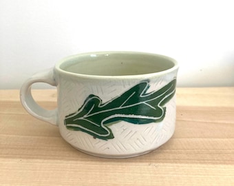 Könnte beinhalten: Handgefertigte Keramik-Tasse mit weißem Hintergrund und grünem Blattmuster. Die Tasse hat einen Henkel und ein strukturiertes Muster auf der Außenseite. Der Innenraum ist hellgrün. Die Tasse steht auf einer Holzoberfläche.