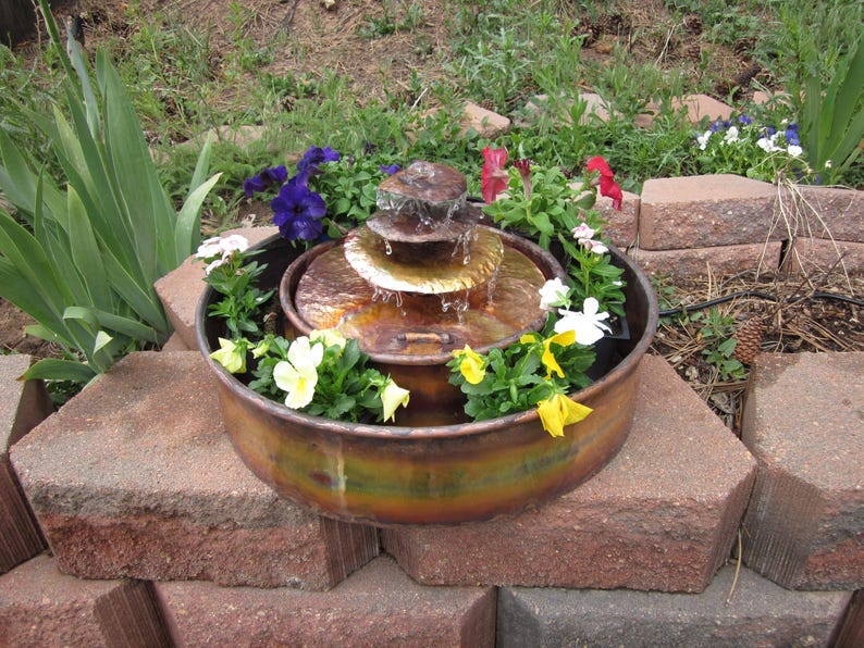 Op de afbeelding: Een drietraps koperen fontein met een waterval, omringd door een verscheidenheid aan kleurrijke bloemen, waaronder paarse, rode, gele en witte.