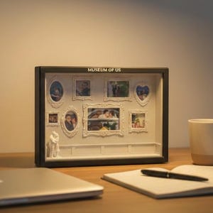 May include: A black shadow box frame, measuring 21cm x 15cm, containing several framed photographs and small figurines. The text "MUSEUM OF US" is printed on the top of the frame. The frame is on a white wall and on a wooden desk.