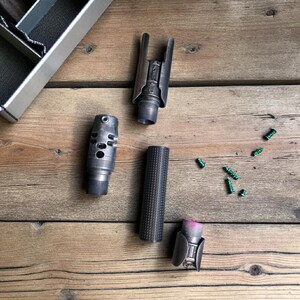 May include: An assortment of metallic gun parts, including a barrel, muzzle brake, and other components, are arranged on a wooden surface. Small green screws are scattered nearby. The parts have a dark, industrial finish.