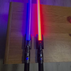 May include: Two toy lightsabers with illuminated blades. One saber glows blue, the other red. Both have black handles with silver accents. The sabers are resting on a wooden surface.