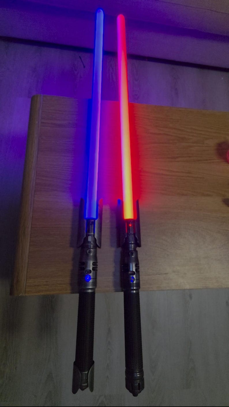 May include: Two toy lightsabers with illuminated blades. One saber glows blue, the other red. Both have black handles with silver accents. The sabers are resting on a wooden surface.