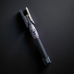 May include: A metallic silver lightsaber prop with a textured black handle. The lightsaber features a detailed design with various cylindrical and rectangular components. The top of the lightsaber has a unique, angular design.