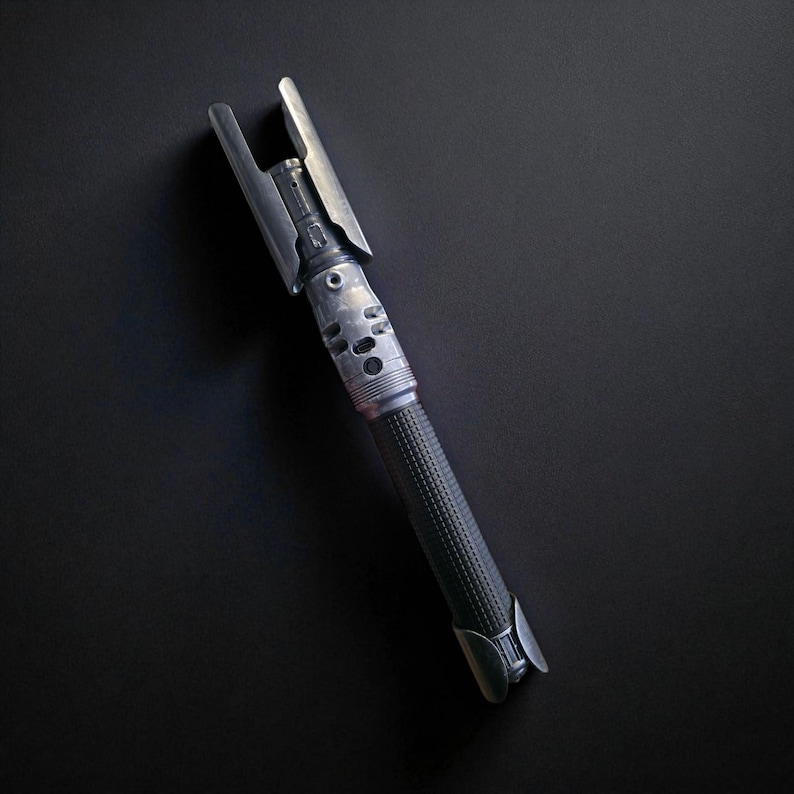May include: A metallic silver lightsaber prop with a textured black handle. The lightsaber features a detailed design with various cylindrical and rectangular components. The top of the lightsaber has a unique, angular design.