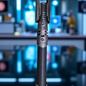 May include: A detailed, metallic lightsaber prop with a textured black handle and silver accents. The hilt features intricate details, including circular and rectangular designs. The top has a distinctive curved guard.