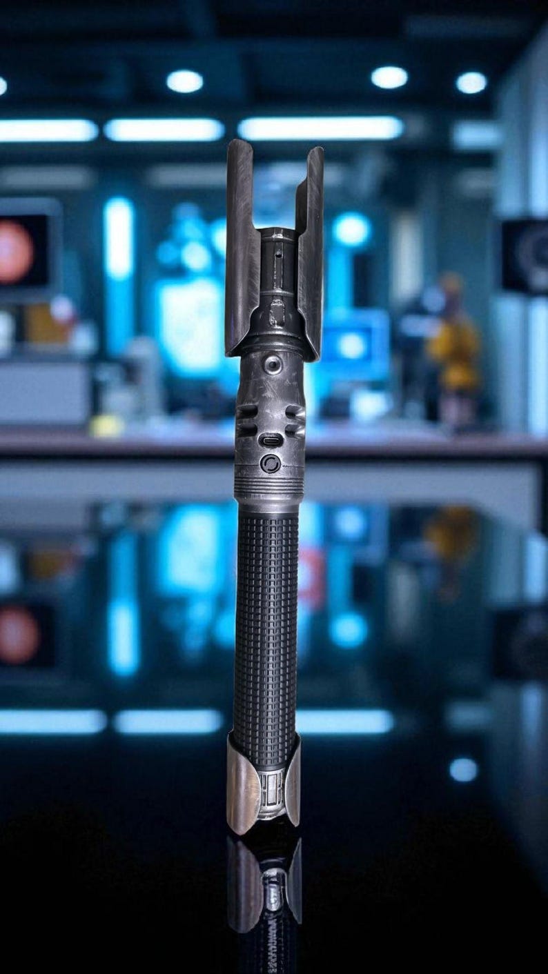 May include: A detailed, metallic lightsaber prop with a textured black handle and silver accents. The hilt features intricate details, including circular and rectangular designs. The top has a distinctive curved guard.