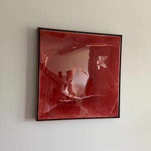 May include: A square wall art piece featuring a deep red, glossy, resin-like surface with a series of raised, irregular, and cracked-looking textures. The artwork is encased in a thin, dark brown frame, creating a reflective and dimensional appearance.
