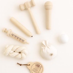 May include: A collection of wooden toys, including a mallet, microphone, syringe, pencil, and a golf ball. Also included is a small white bag with a flower and a wooden tag with a golf-themed design. The toys are arranged on a white surface.