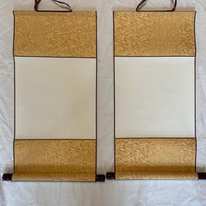 Könnte beinhalten: Zwei leere Schriftrollen mit einem cremefarbenen Papierzentrum und gold gemusterten oberen und unteren Abschnitten. Die Schriftrollen haben dunkelbraune Holzdübel und Aufhängeschnüre.
