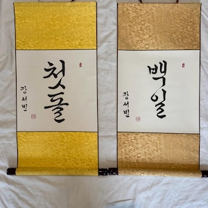 May include: Two Korean calligraphy scrolls with brown leather hanging straps. The scrolls feature gold and cream-colored panels with black calligraphy. The left scroll has the text "첫돌" and the right scroll has the text "백일".