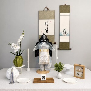 Könnte beinhalten: Dekoration für eine traditionelle koreanische Feier zum ersten Geburtstag. Mit einer Miniatur-Tracht, Schriftrollen, Kerzen und dekorativen Gegenständen auf einer weißen Tischdecke. Enthält eine Vase mit weißen Blumen und einen kleinen Holzrahmen.