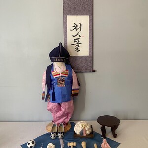 May include: A traditional Korean hanbok set for a child, including a blue vest, pink trousers, and a dark blue hat. A wall scroll with Korean calligraphy hangs above. Accessories include white shoes, a small wooden table, and various small items.