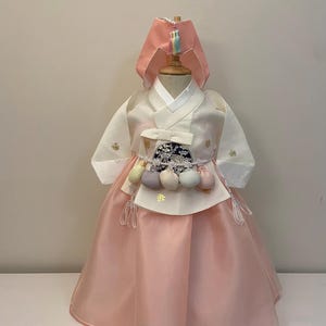 May include: A traditional Korean Hanbok dress for a child. The dress features a white top with long sleeves and a pink skirt. The outfit includes a matching pink headpiece and decorative accents.