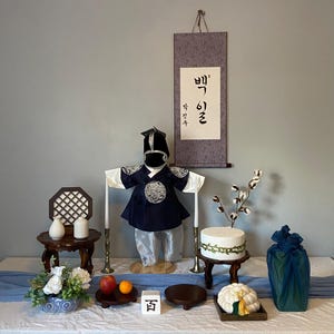 May include: A Korean baby's first birthday display featuring a navy and white outfit and hat. The scene includes a scroll with Korean text, a small table, candles, fruit, and a cake. The table is set with various traditional items.