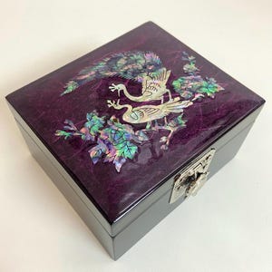 Può includere: Un portagioie quadrato, viola scuro, con finitura lucida. Il coperchio presenta un disegno intarsiato di due uccelli e fogliame in madreperla iridescente. La scatola ha una chiusura color argento.