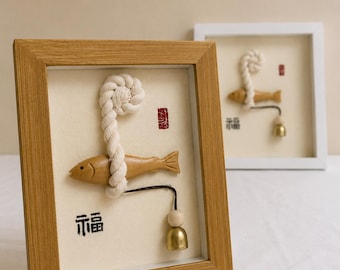 May include: A framed wall art piece featuring a light wood frame and a cream-colored background. A carved wooden fish is wrapped in a thick white rope, accompanied by a small brass bell on a dark cord and the black Chinese character for fortune.