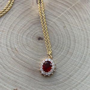 May include: A gold chain necklace with a red oval gemstone pendant surrounded by smaller clear gemstones.