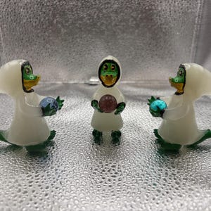 Pode incluir: Três figuras fantásticas de criaturas de rosto verde em vestes brancas com capuz. Cada uma segura um orbe colorido. As figuras têm caudas verdes e bicos amarelos. O fundo é uma superfície prateada texturizada.