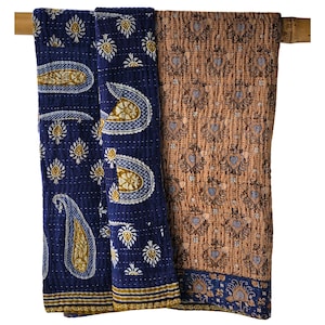 May include: A reversible quilted throw blanket featuring a deep blue side with white and gold paisley and floral patterns, and a tan side with a repeating heart-shaped motif. The fabric displays visible running stitch details throughout the surface.