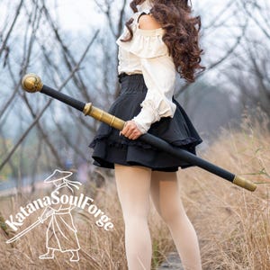 May include: A black and gold staff with a spherical top and decorative gold bands. The staff is held by a person wearing a white blouse and black skirt. The text "Katana SoulForge" is visible in the lower left corner.