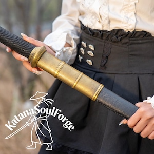 May include: A close-up of a katana sword with a black handle and a gold-colored hilt. The sword is held by a person. The Katana SoulForge logo is in the lower left corner.