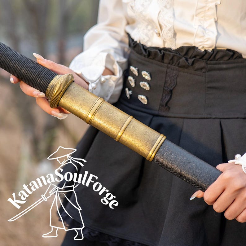 May include: A close-up of a katana sword with a black handle and a gold-colored hilt. The sword is held by a person. The Katana SoulForge logo is in the lower left corner.