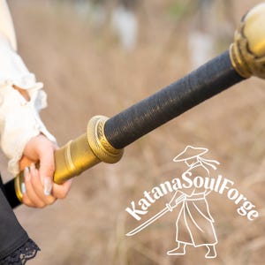 May include: A close-up of a sword hilt and handle. The hilt is gold-colored with intricate detailing, and the handle is wrapped in black material. The text "Katana SoulForge" is visible.