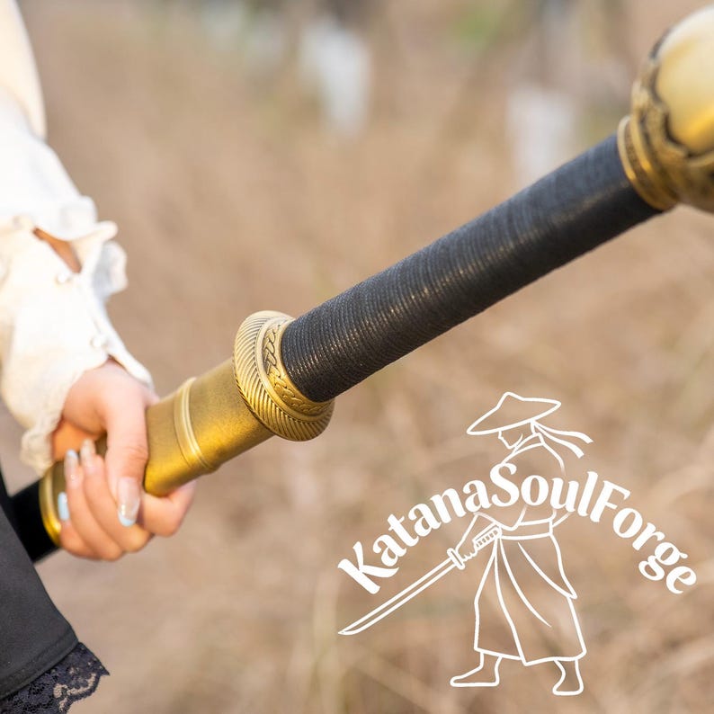 May include: A close-up of a sword hilt and handle. The hilt is gold-colored with intricate detailing, and the handle is wrapped in black material. The text "Katana SoulForge" is visible.