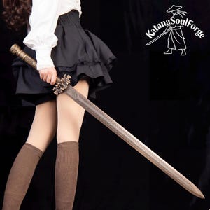 May include: A long, ornate sword with a bronze-colored hilt and a silver-colored blade. The blade has a detailed etched design. The sword is held by a person wearing a white blouse, black skirt, and brown knee-high socks. The Katana SoulForge logo is in the upper right corner.