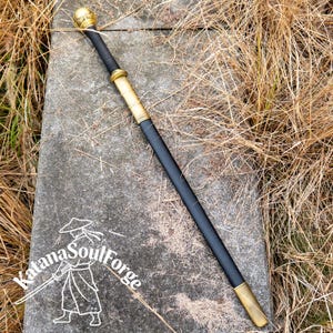 May include: A black walking stick with gold-colored accents and a rounded top. The stick has a gold-colored band near the top and bottom. The text "Katana SoulForge" is visible in the lower left corner.